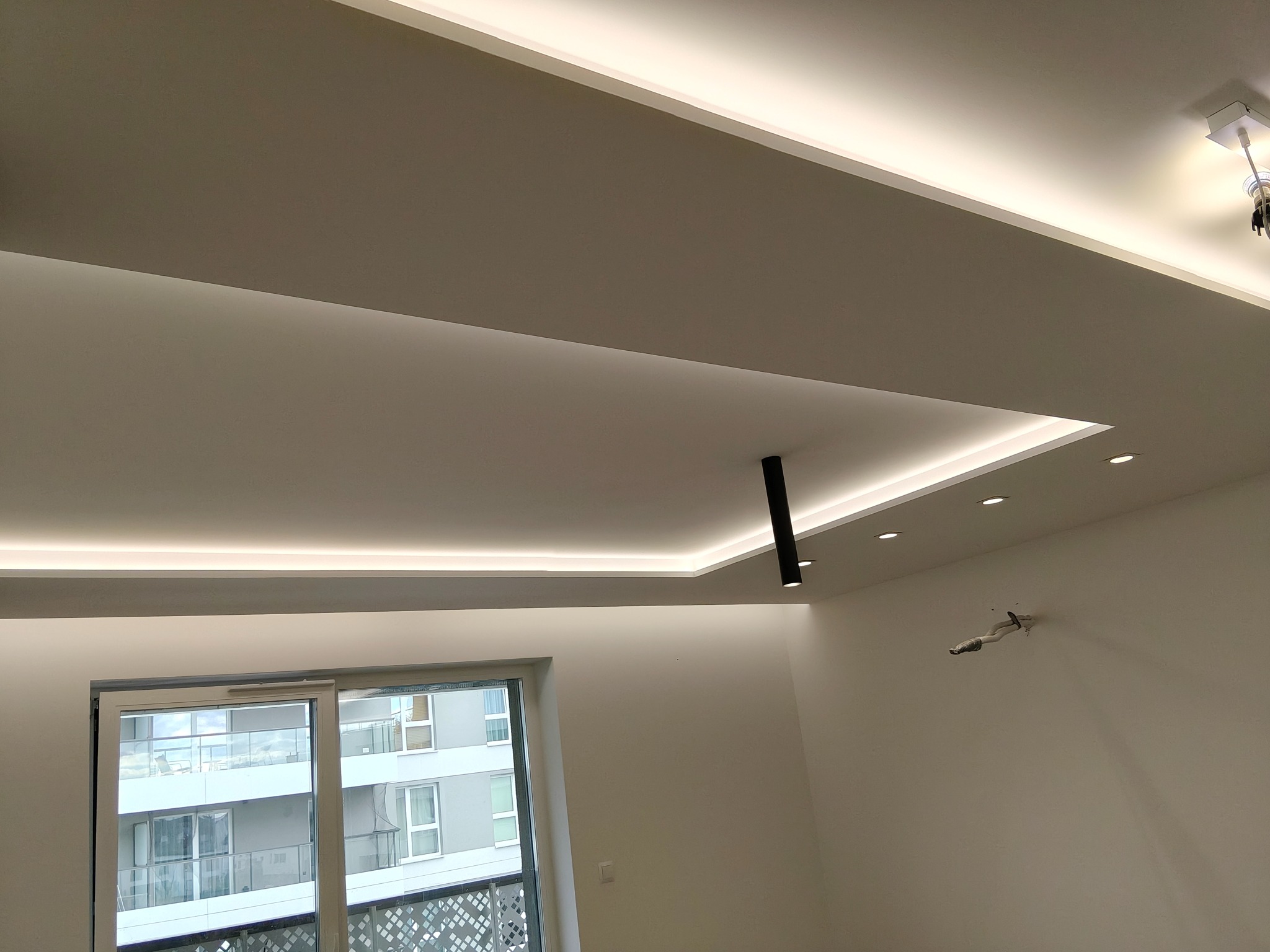 Nowoczesny sufit podwieszany z oświetleniem LED, czarna lampa punktowa i okno z widokiem na budynek mieszkalny. Minimalistyczny design wnętrza.