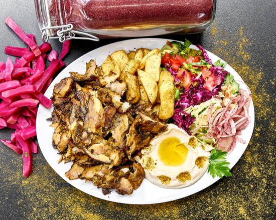 Talerz z shawarmą z kurczaka, frytkami, hummusem z oliwą i sałatką warzywną, uzupełniony różową marynowaną rzodkiewką i posypany przyprawami, w tle słoik z czerwoną przyprawą.