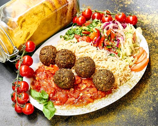 Talíř s falafelovými kuličkami na rajčatovém základu, rýží, zeleninovým salátem a rajčátky, vedle sklenice s kořením, posypané kořením.