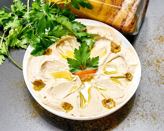 Kremowy hummus na białym talerzu, udekorowany świeżą natką pietruszki, papryką i polany oliwą, z przyprawami rozsypanymi wokół talerza i słoikiem przypraw w tle.