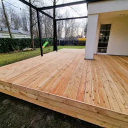 Nowoczesny taras z jasnego drewna z czarną konstrukcją pergoli w Warszawie, widok na trawnik i dom z żółtym busem w tle.