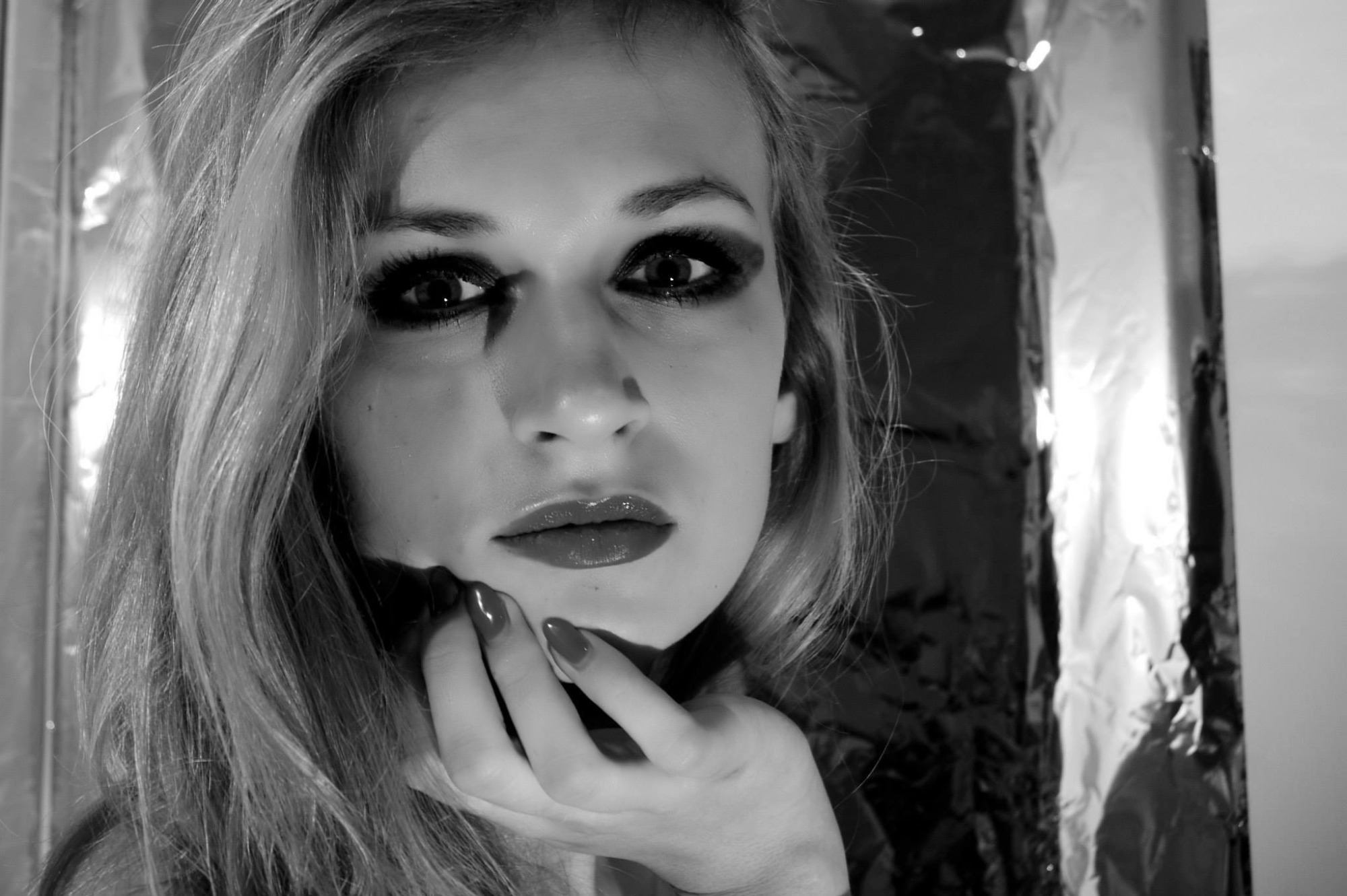 Portret kobiety z mocnym makijażem oczu i ustami w odcieniu czerwieni, opierającej brodę na dłoni, w czarno-białej tonacji na tle folii.