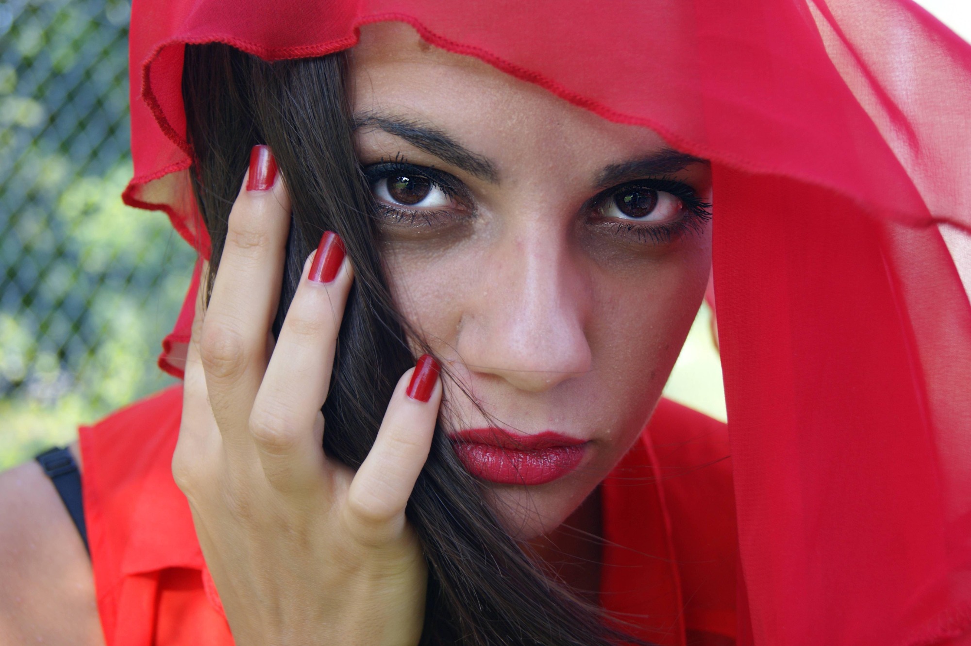 Portret kobiety z ciemnymi włosami i czerwonym szalem, z intensywnym spojrzeniem w obiektyw i czerwonymi paznokciami.