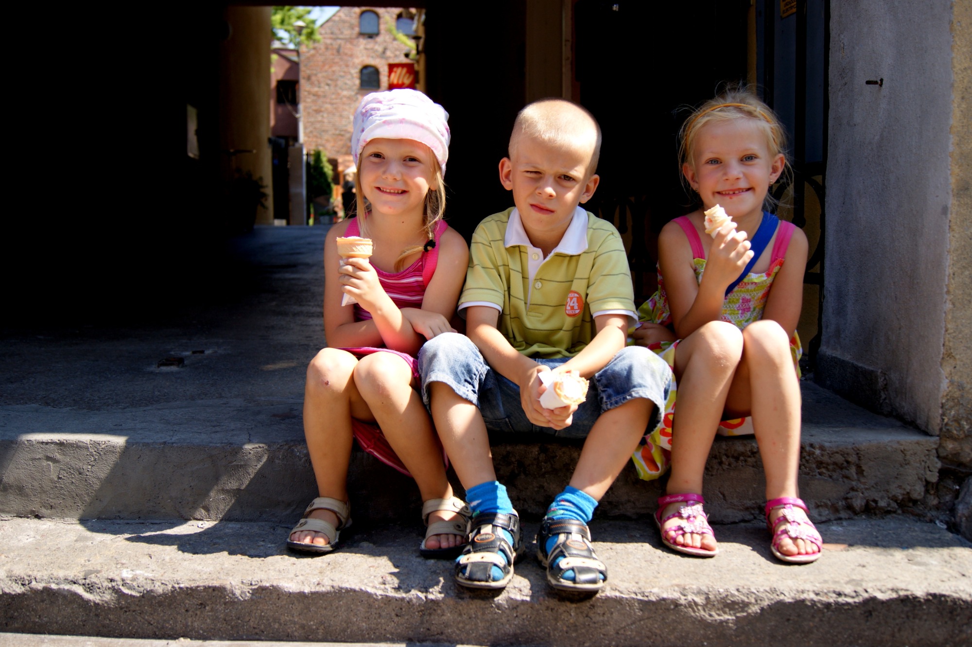 Troje uśmiechniętych dzieci siedzi na betonowych schodach w cieniu, każde trzyma loda w wafelku, w tle widoczny budynek z czerwonej cegły.