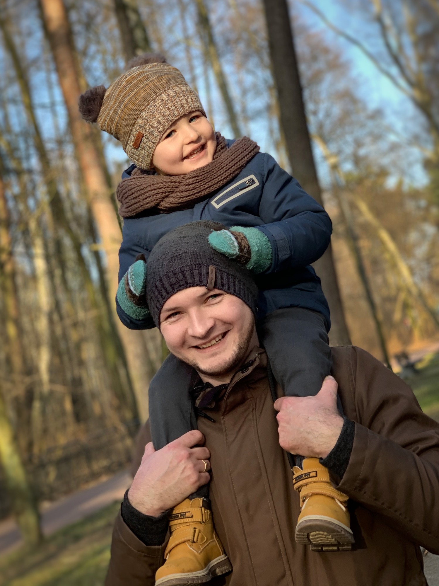 Uśmiechnięty ojciec trzyma na ramionach małego synka w zimowej scenerii leśnej, obaj w czapkach i kurtkach, z wyraźnym bokeh w tle.