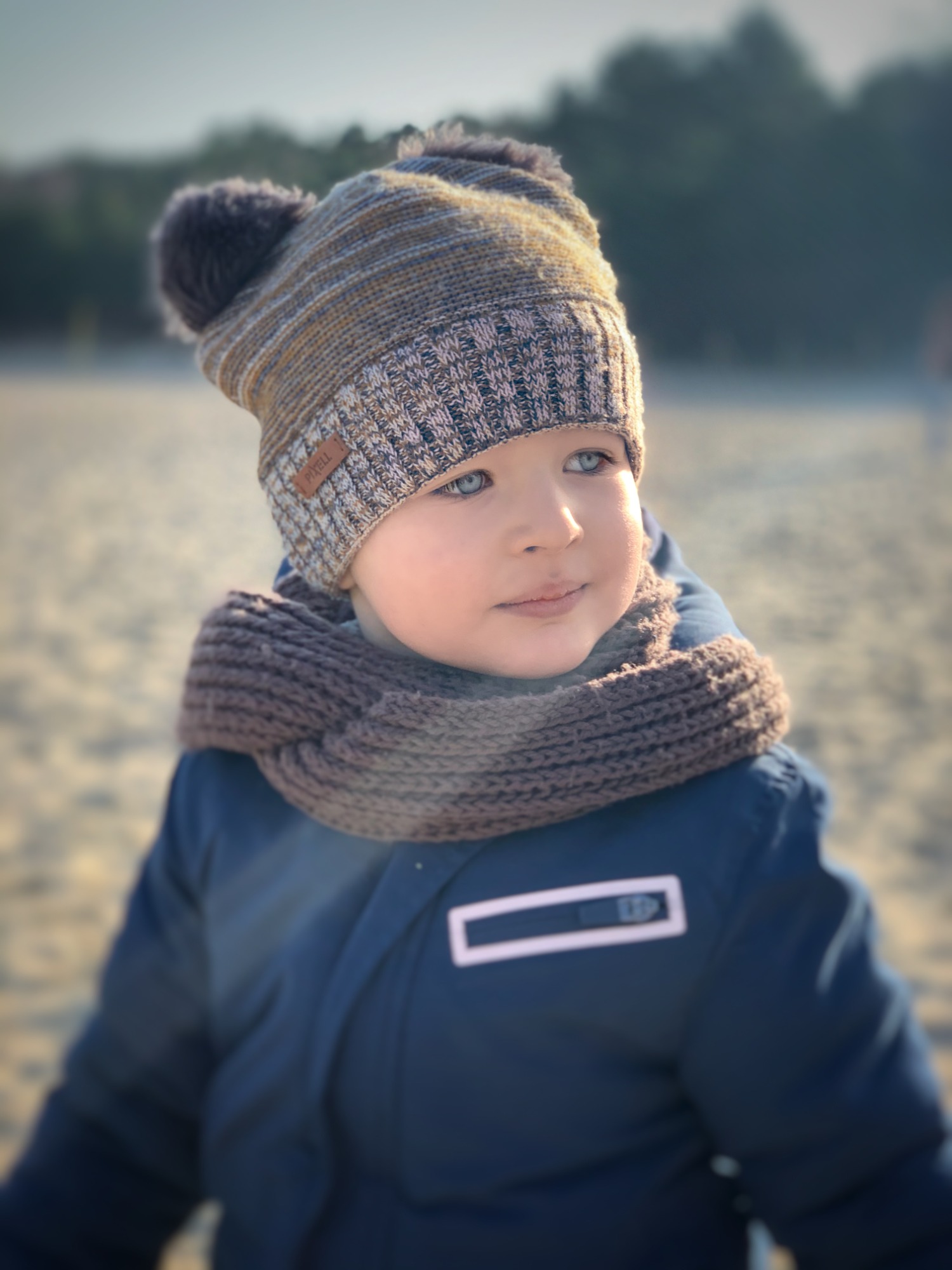 Portret małego chłopca w czapce z pomponami i grubym szaliku, zamyślony wzrok skierowany w bok, na rozmytym tle piaszczysta plaża i zieleń.