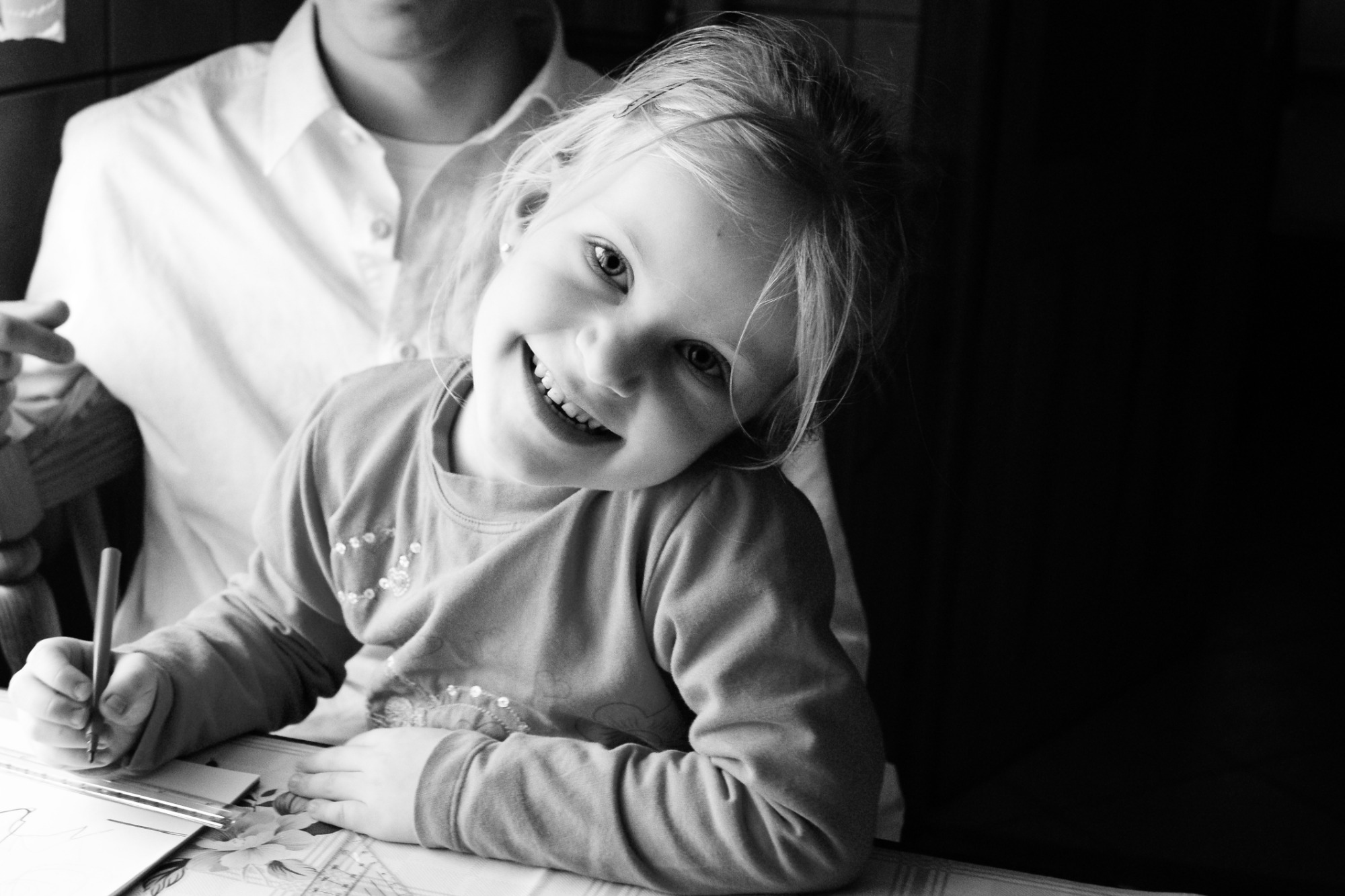 Czarno-białe zdjęcie uśmiechniętej dziewczynki rysującej przy stole, z widocznym fragmentem osoby dorosłej w tle.