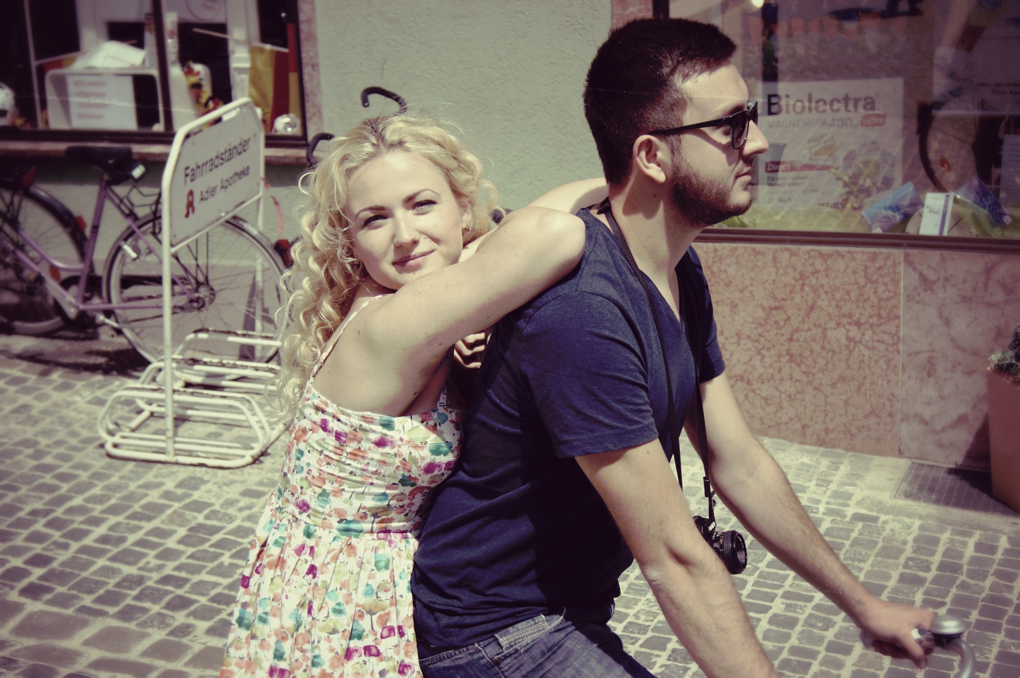 Para na rowerze na tle brukowanej ulicy i witryny sklepowej, kobieta w kwiecistej sukience obejmuje mężczyznę w okularach.