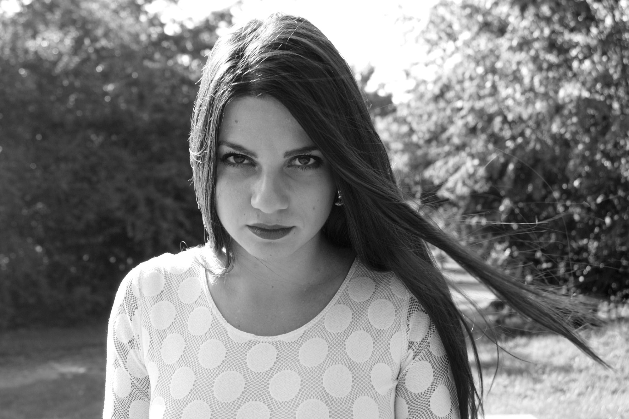 Portret kobiety z długimi, rozwianymi włosami na tle rozmytej zieleni, w białej sukience w grochy, czarno-białe zdjęcie.