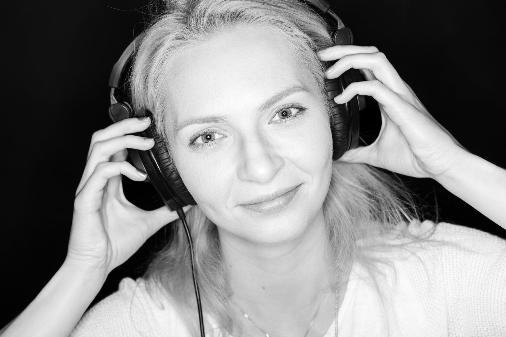 Portret kobiety w czarno-białej tonacji, uśmiechniętej, z słuchawkami na uszach, trzymającej je dłońmi, na ciemnym tle.