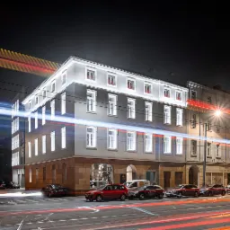 Odrestaurowana kamienica nocą, z jasnymi oknami i subtelnym oświetleniem elewacji, z długimi smugami świateł samochodowych na pierwszym planie.