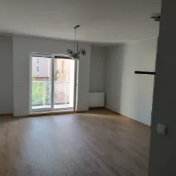 Pusty, jasny pokój z drewnianą podłogą, widok na balkon i nowoczesną lampę wiszącą z szarymi kulami.