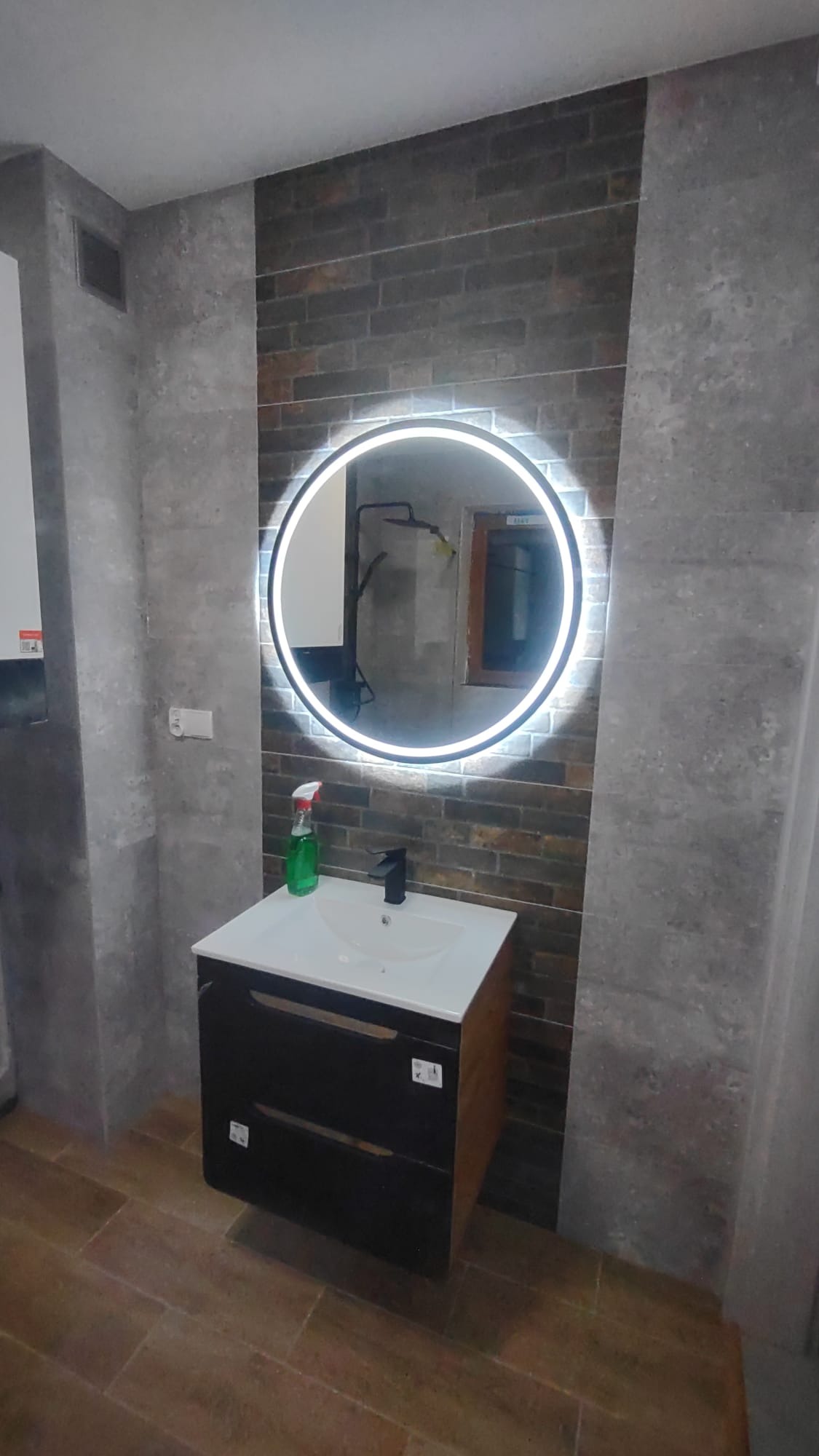 Ukończona łazienka: ciemna szafka z umywalką, okrągłe lustro z podświetleniem LED, płytki imitujące cegłę i szare płytki na ścianach, brązowa podłoga.