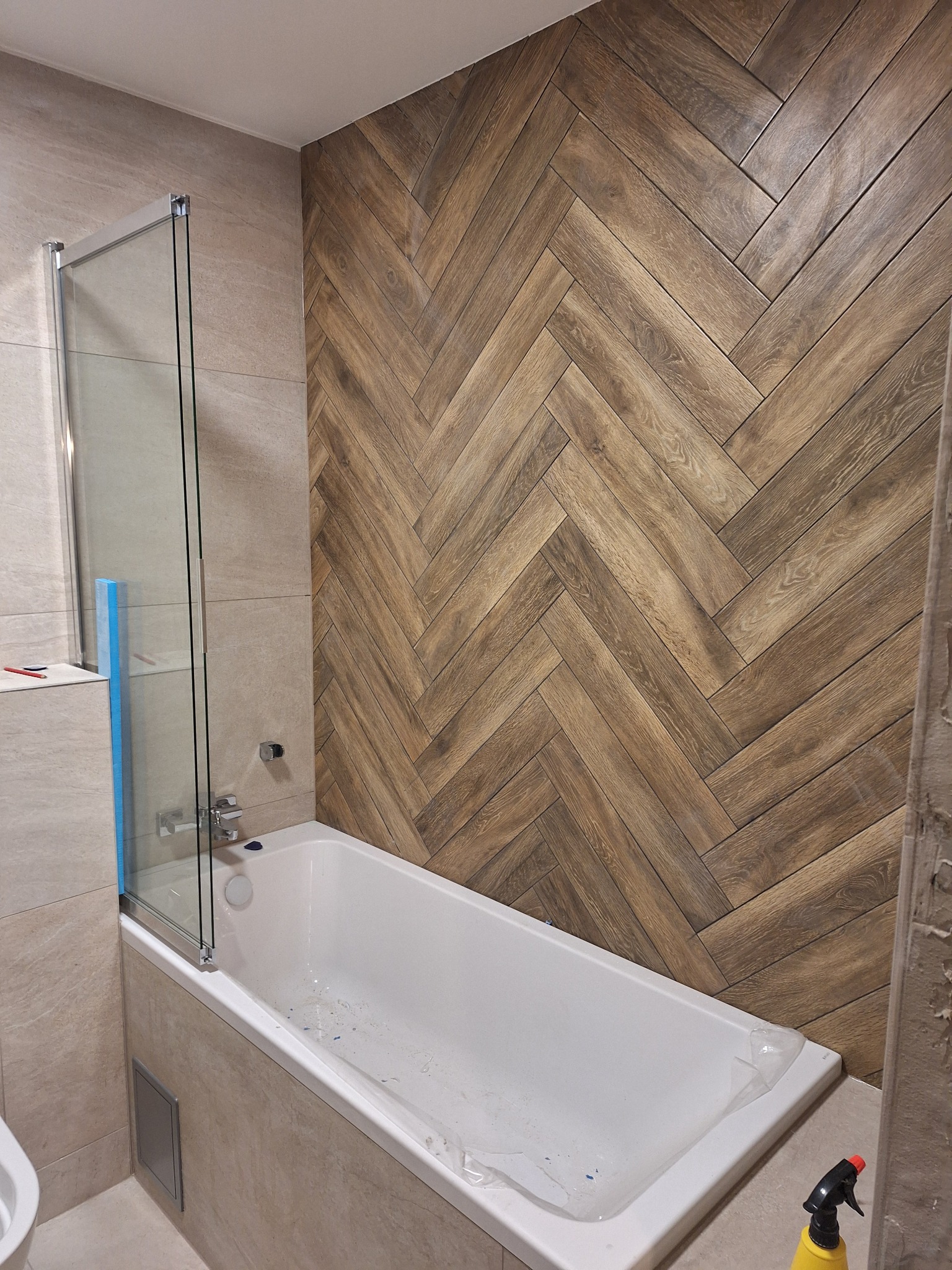 Wykończona łazienka w Lubiczu Górnym: ściana z płytek imitujących drewno ułożonych w jodełkę, biała wanna z parawanem nawannowym i beżowe płytki na pozostałych ścianach.