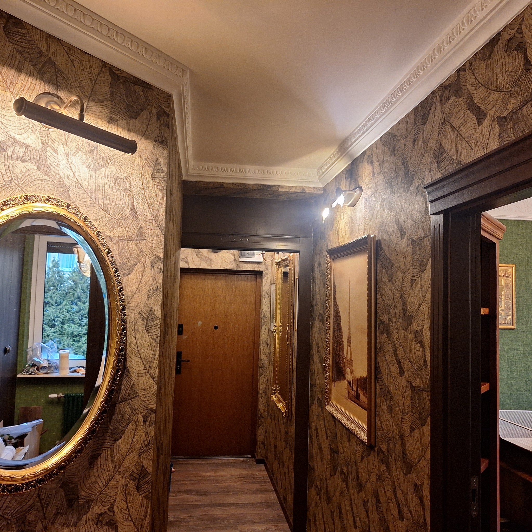 Korytarz z tapetą w liście, ozdobnym lustrem w złotej ramie i obrazem z wieżą Eiffla. Dekoracyjne listwy sufitowe. Widok na drzwi wejściowe i inne pomieszczenia.