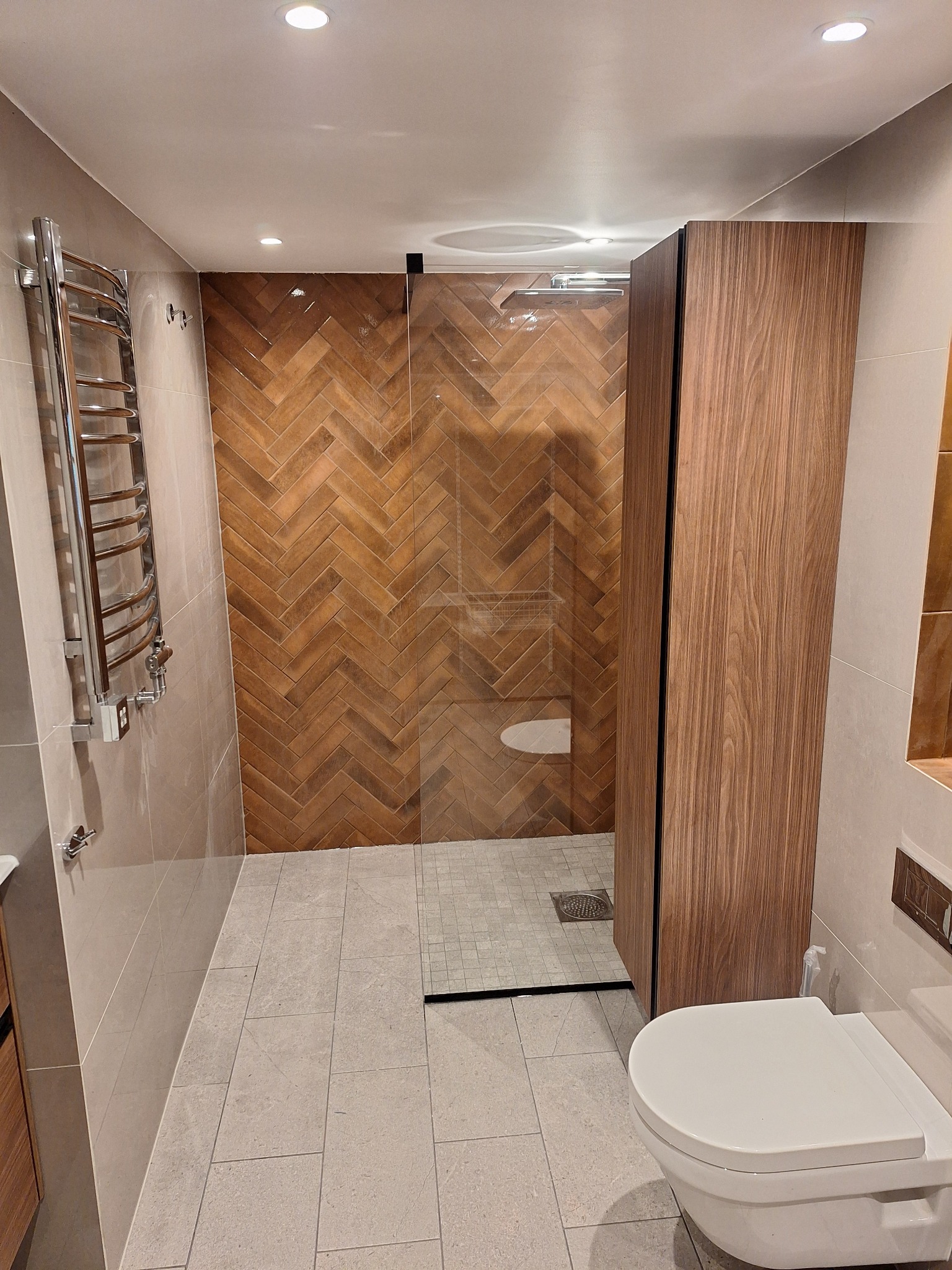 Nowoczesna łazienka z kabiną prysznicową typu walk-in, wykończona płytkami w jodełkę. Widoczny grzejnik drabinkowy i podwieszana toaleta. Minimalistyczny design.