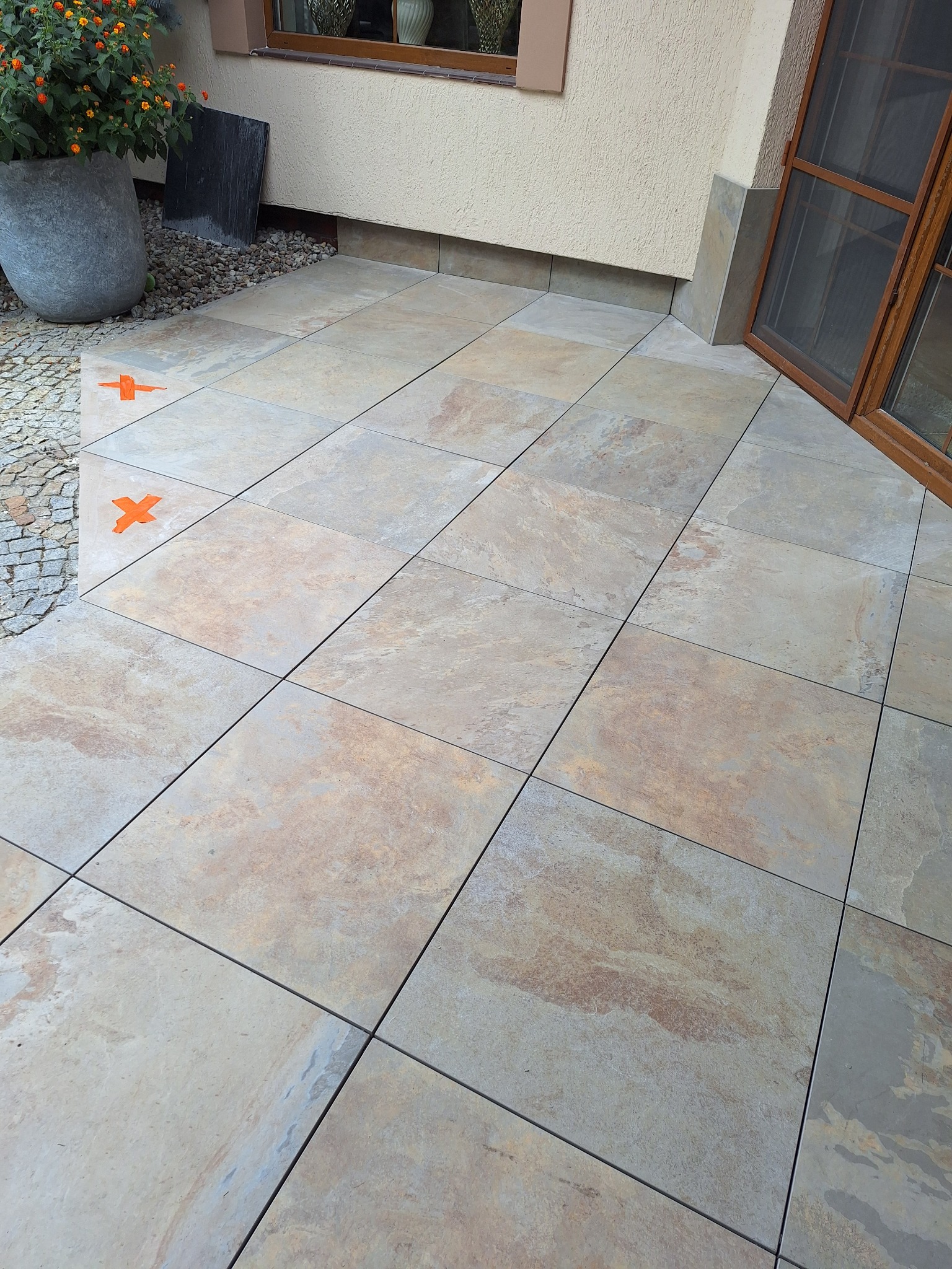 Ułożona, beżowo-brązowa, kwadratowa terakota na zewnątrz budynku. Widoczne krzyżyki dystansowe, donica z kwiatami i fragment okna.