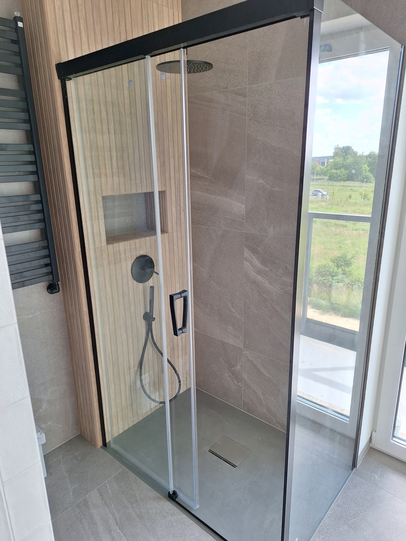 Nowoczesna kabina prysznicowa z czarnymi profilami i deszczownicą, wykończona płytkami w odcieniach beżu i szarości. Widok na zewnątrz przez okno.
