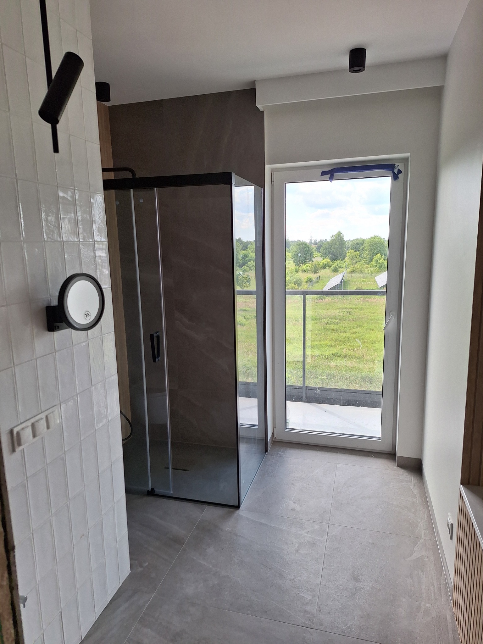 Nowoczesna łazienka z kabiną prysznicową typu walk-in, szarą podłogą z płytek i dużym oknem z widokiem na zieleń. Minimalistyczne oświetlenie i lustro dodają elegancji.