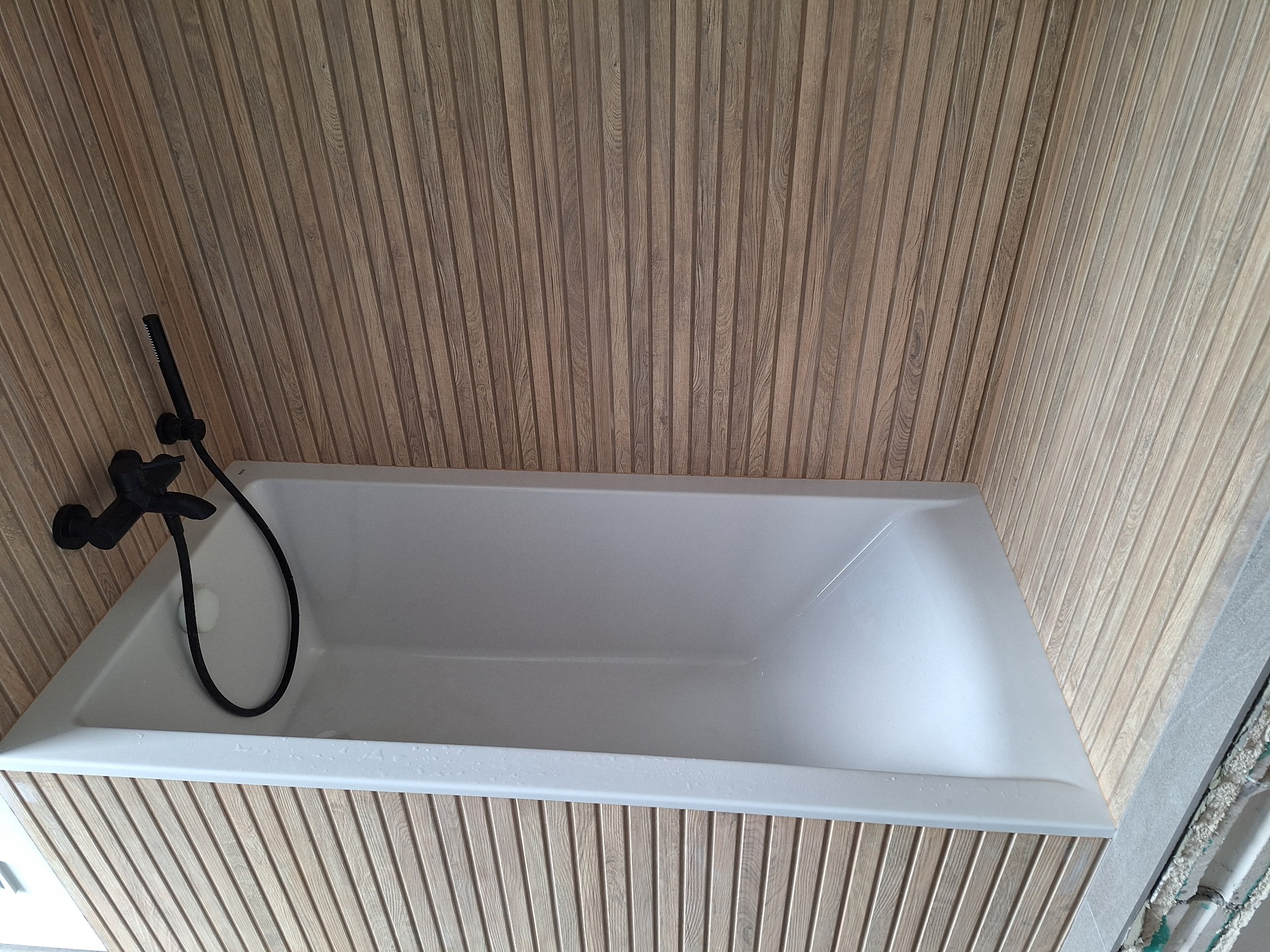 Nowoczesna łazienka z wanną i armaturą w kolorze czarnym, wykończona panelami ściennymi imitującymi drewno. Minimalistyczny design, ciepłe oświetlenie.