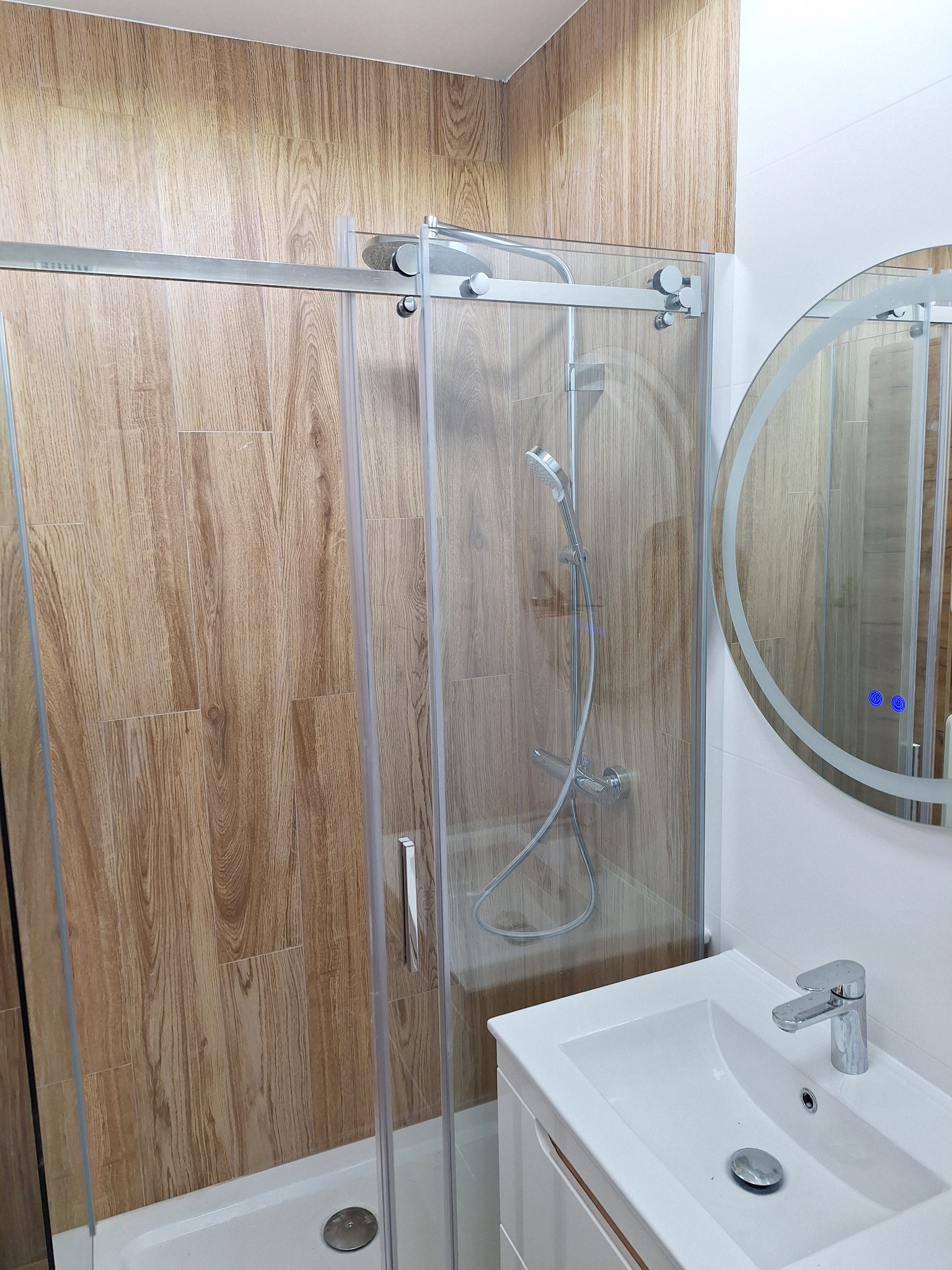 Nowoczesna kabina prysznicowa z przesuwnymi drzwiami, drewnopodobne płytki na ścianie, okrągłe lustro z podświetleniem i biała umywalka z szafką.