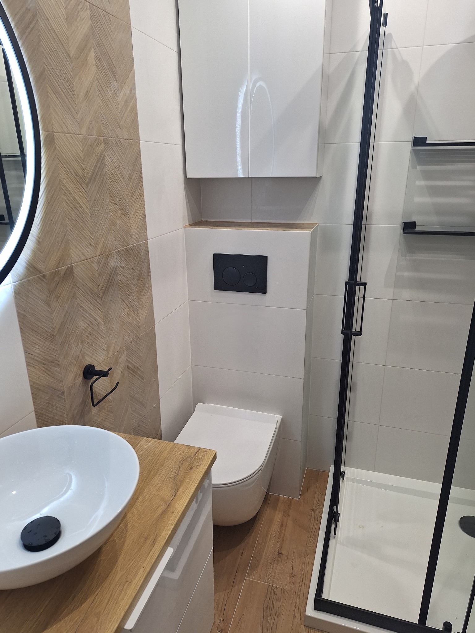 Nowoczesna łazienka z umywalką nablatową, kabiną prysznicową z czarnymi profilami i toaletą podwieszaną. Na ścianie dekoracyjne płytki imitujące drewno.