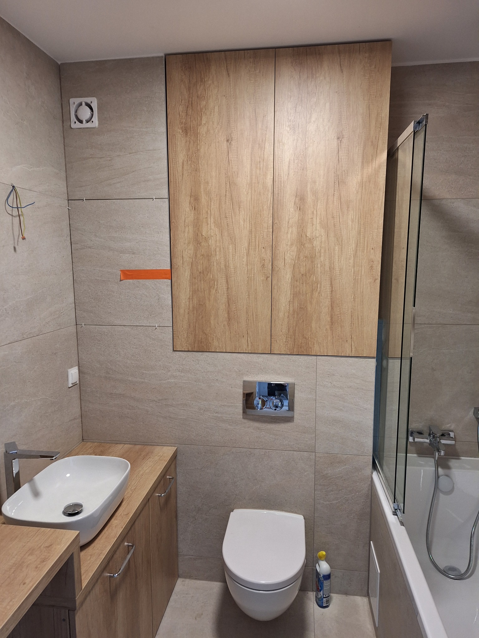 Jasna łazienka z beżowymi płytkami, drewnianymi szafkami i białym wyposażeniem: umywalką, toaletą i wanną z parawanem. Widoczny wentylator i butelka płynu.