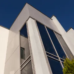 Elewacja budynku z dużymi, ciemnymi oknami pod kątem, z widocznym odbiciem innego okna i fragmentem iglastego krzewu na pierwszym planie, na tle błękitnego nieba.