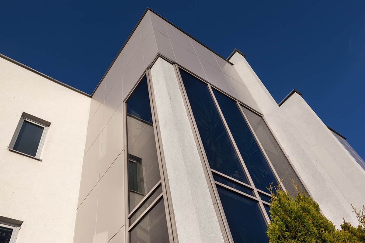 Elewacja budynku z dużymi, ciemnymi oknami pod kątem, z widocznym odbiciem innego okna i fragmentem iglastego krzewu na pierwszym planie, na tle błękitnego nieba.