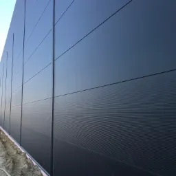 Ściana budynku przemysłowego pokryta ciemnoszarymi płytami warstwowymi z widoczną strukturą poziomych rowków, z przygotowanym podłożem gruntowym u dołu, sfotografowana pod kątem z boku.