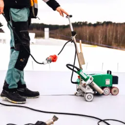 Pracownik w roboczych spodniach łączy elektrycznie zgrzewarkę do membran dachowych na płaskim dachu, w tle widoczne drzewa.