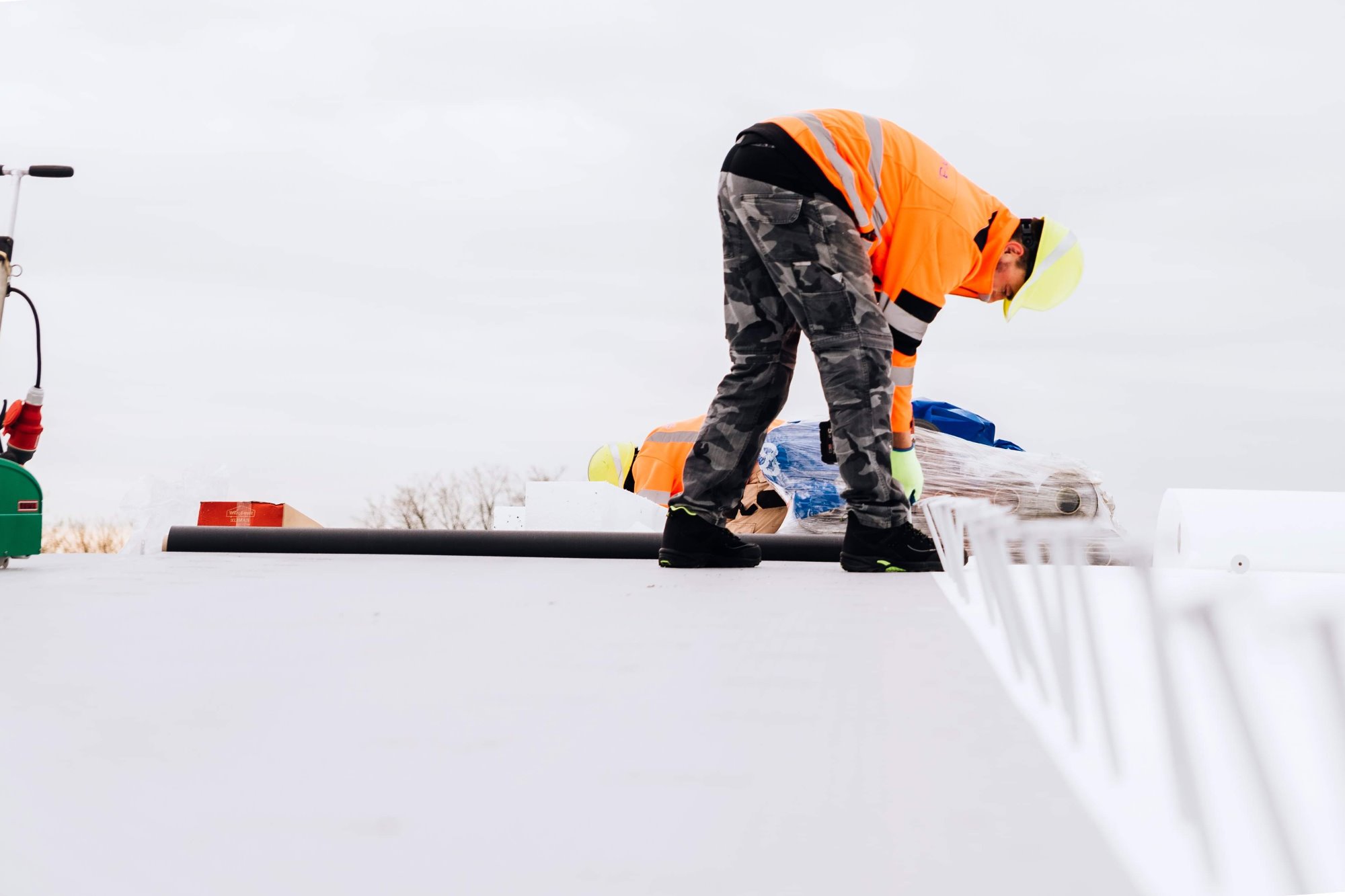 Pracownik w odblaskowej kurtce i kasku przygotowuje materiały izolacyjne na białym dachu płaskim, widoczne rolki folii i narzędzia w tle.