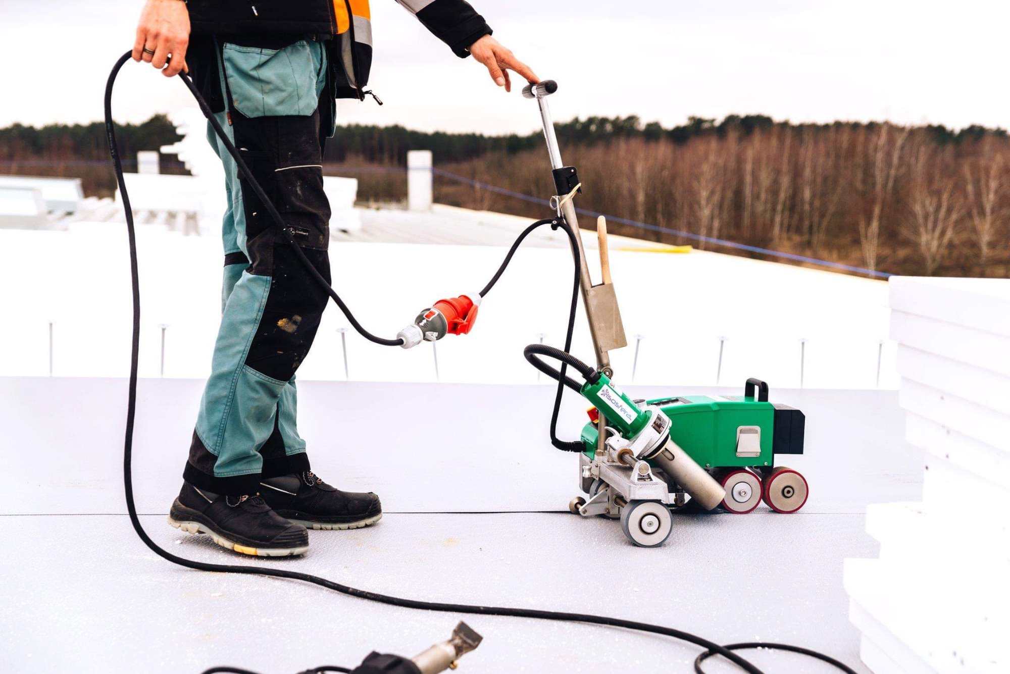 Pracownik w roboczych spodniach łączy elektrycznie zgrzewarkę do membran dachowych na płaskim dachu, w tle widoczne drzewa.