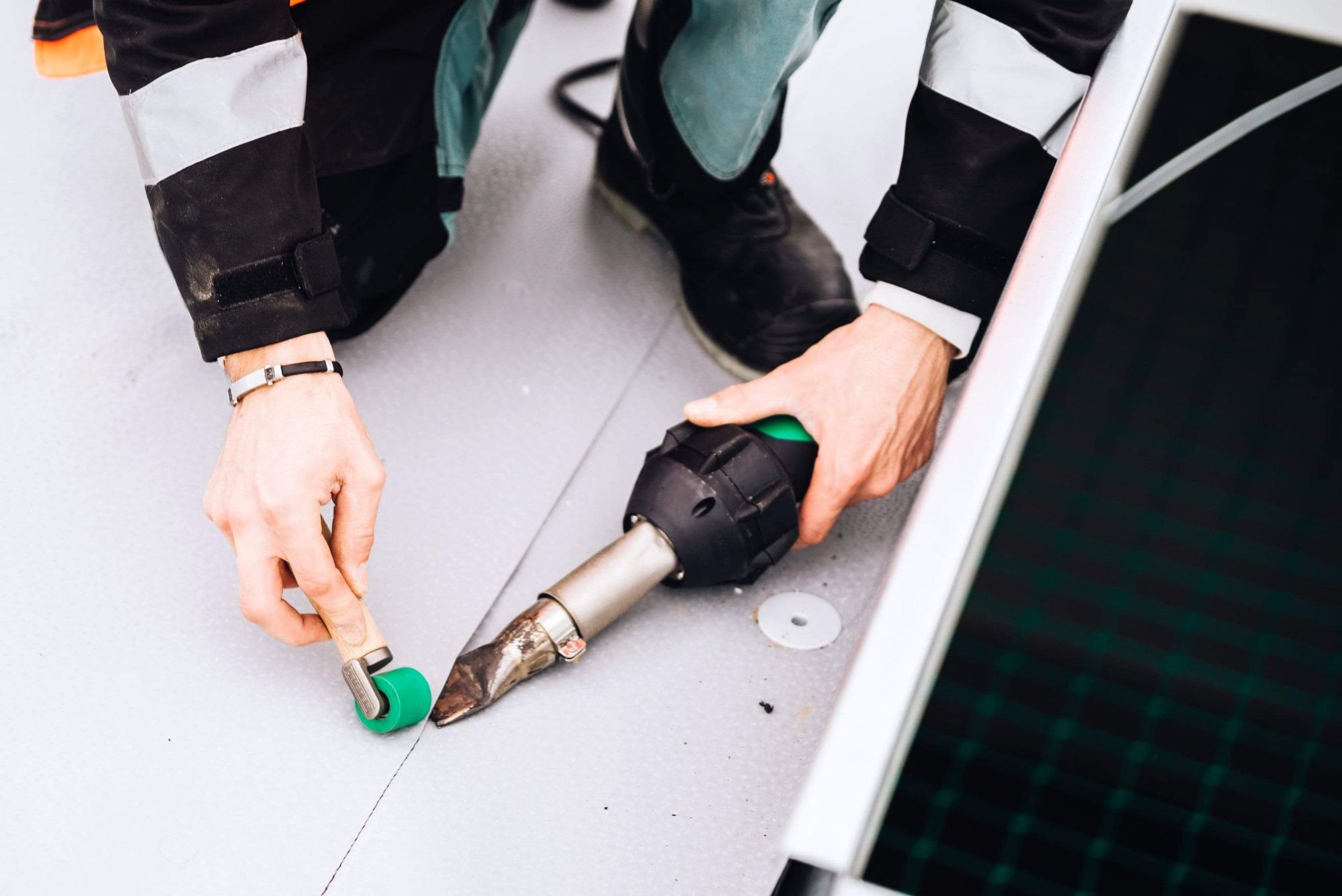 Pracownik łączy płyty warstwowe za pomocą zgrzewarki i zielonego wałka dociskowego na jasnoszarym podłożu, widoczne elementy stroju roboczego i fragment czarnej płyty.