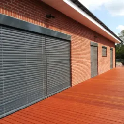 Ceglany dom z szarymi żaluzjami zewnętrznymi i drewnianym tarasem w słoneczny dzień.