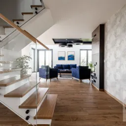 Nowoczesny salon z drewnianą podłogą, szklanymi schodami i niebieskimi meblami wypoczynkowymi, jasne wnętrze domu w Krakowie z akcentami drewna i roślinności.