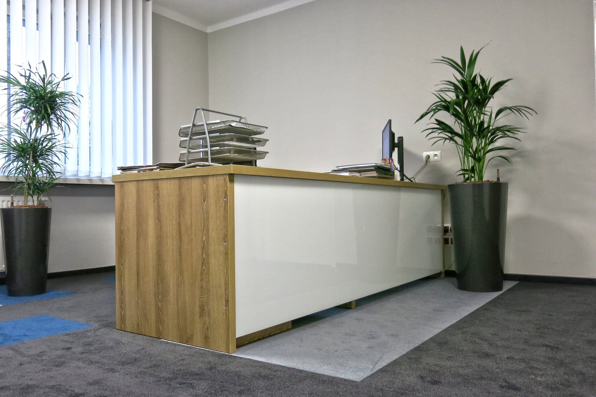 Nowoczesna recepcja w biurze z drewnianymi elementami, białym frontem, metalowymi organizerami na dokumenty i doniczkowymi roślinami po bokach.