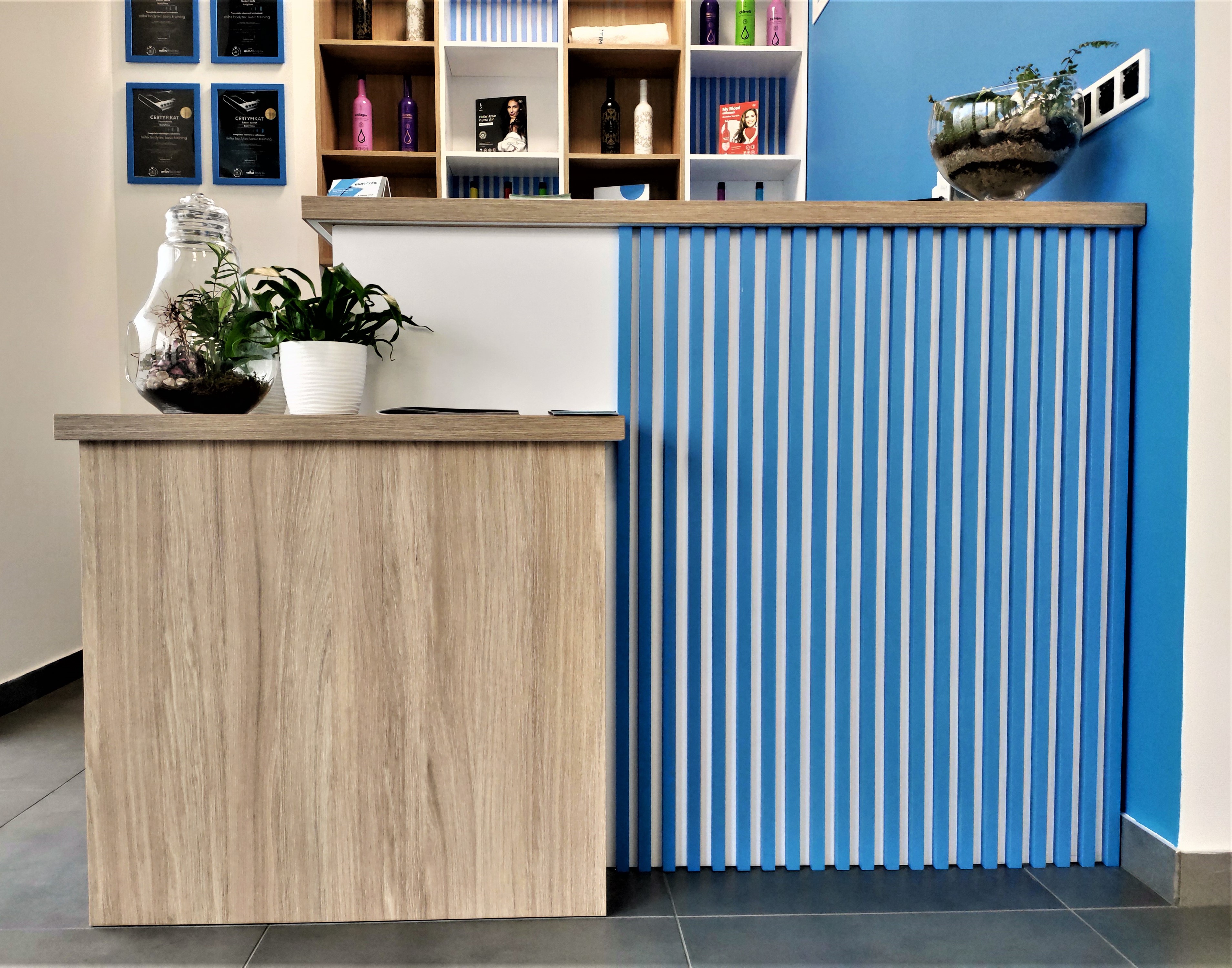 Recepcja w salonie BodyTime w Krakowie z drewnianym blatem, niebieskimi pionowymi panelami, roślinami w szklanych naczyniach i certyfikatami na ścianie.