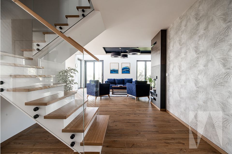 Nowoczesny salon z drewnianą podłogą, szklanymi schodami i niebieskimi meblami wypoczynkowymi, jasne wnętrze domu w Krakowie z akcentami drewna i roślinności.