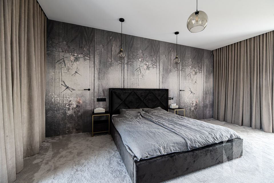 Nowoczesna sypialnia z ciemnoszarym łóżkiem z welurowym zagłówkiem, nocnymi szafkami z metalowymi nóżkami i tapetą w leśny motyw z ukrytymi drzwiami.