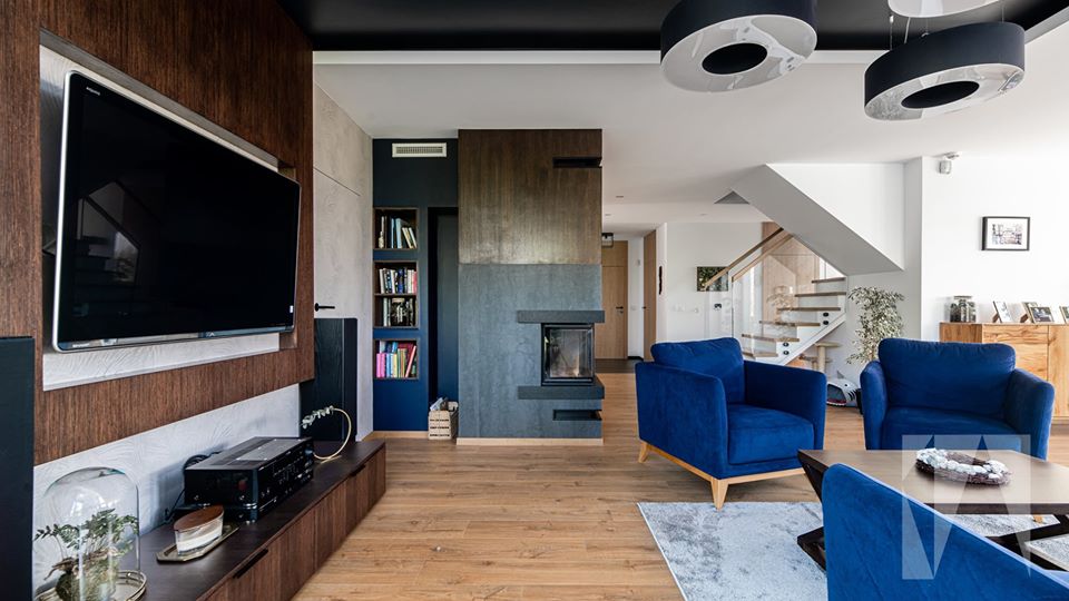 Nowoczesny salon w domu w Krakowie: drewniana podłoga, niebieskie fotele, kominek z szarego kamienia, zabudowa TV z drewnianymi panelami i czarne, okrągłe lampy sufitowe.