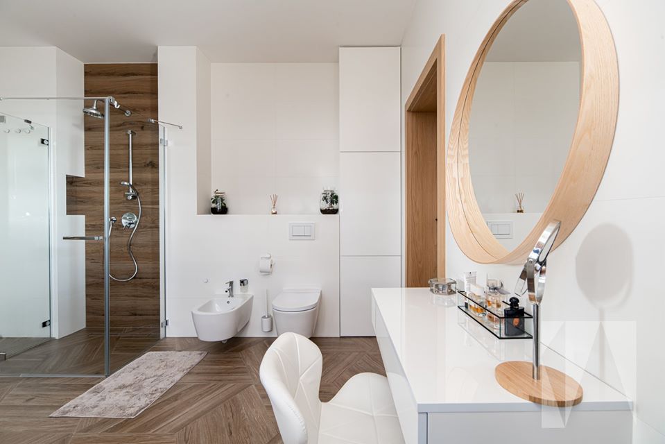 Nowoczesna łazienka z drewnianymi akcentami: kabina prysznicowa z brązową ścianą, biała toaleta i bidet, okrągłe lustro nad białą toaletką, brązowa podłoga w jodełkę.