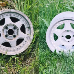 Dwa aluminiowe felgi samochodowe na trawie: jeden po piaskowaniu z widocznymi śladami użytkowania, drugi po malowaniu proszkowym na jasny kolor.