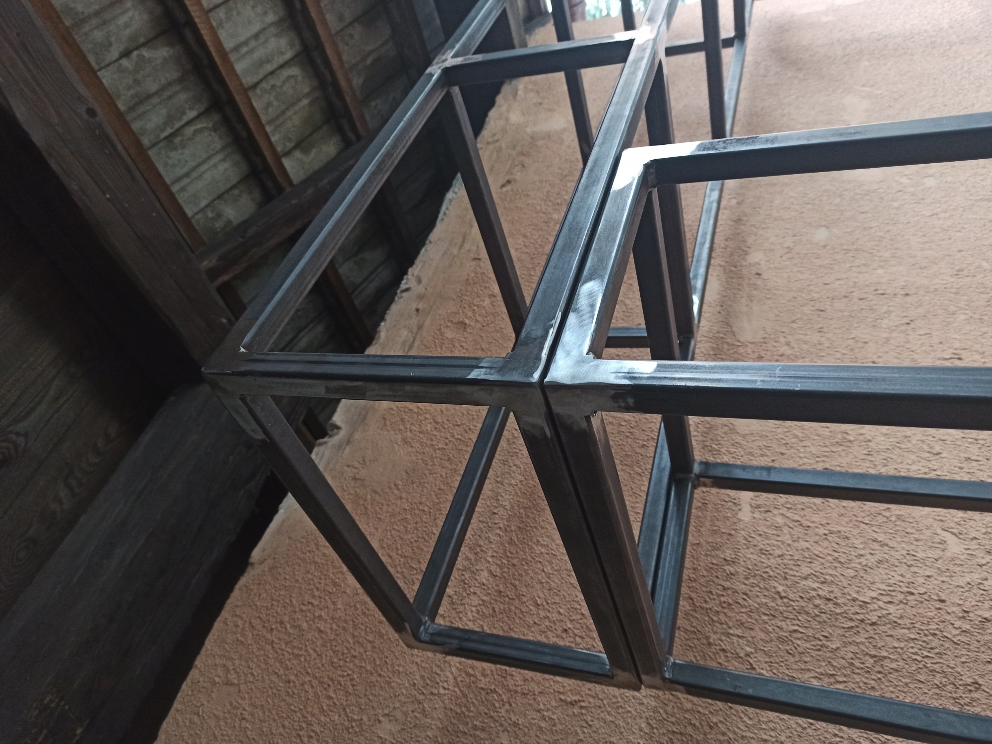 Metalowa konstrukcja szkieletowa z profili zamkniętych, spawana, widoczne spoiny, surowy metal, na tle drewnianego stropu i ściany o chropowatej fakturze.