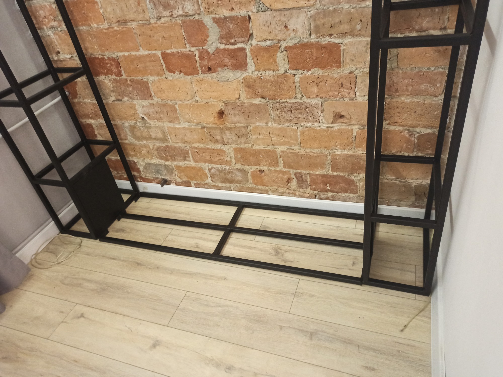 Metalowa konstrukcja regału o czarnym, matowym wykończeniu, spawana z profili, na tle ceglanej ściany i jasnej podłogi z paneli.