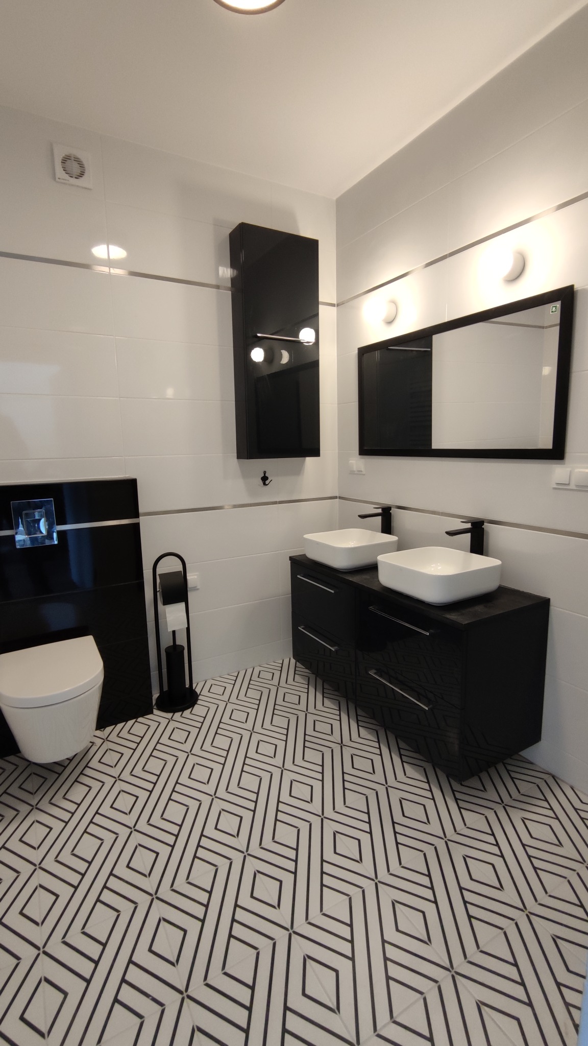Nowoczesna łazienka z białymi płytkami, czarnymi meblami i geometryczną podłogą, oświetlenie punktowe nad lustrem i wisząca szafka.