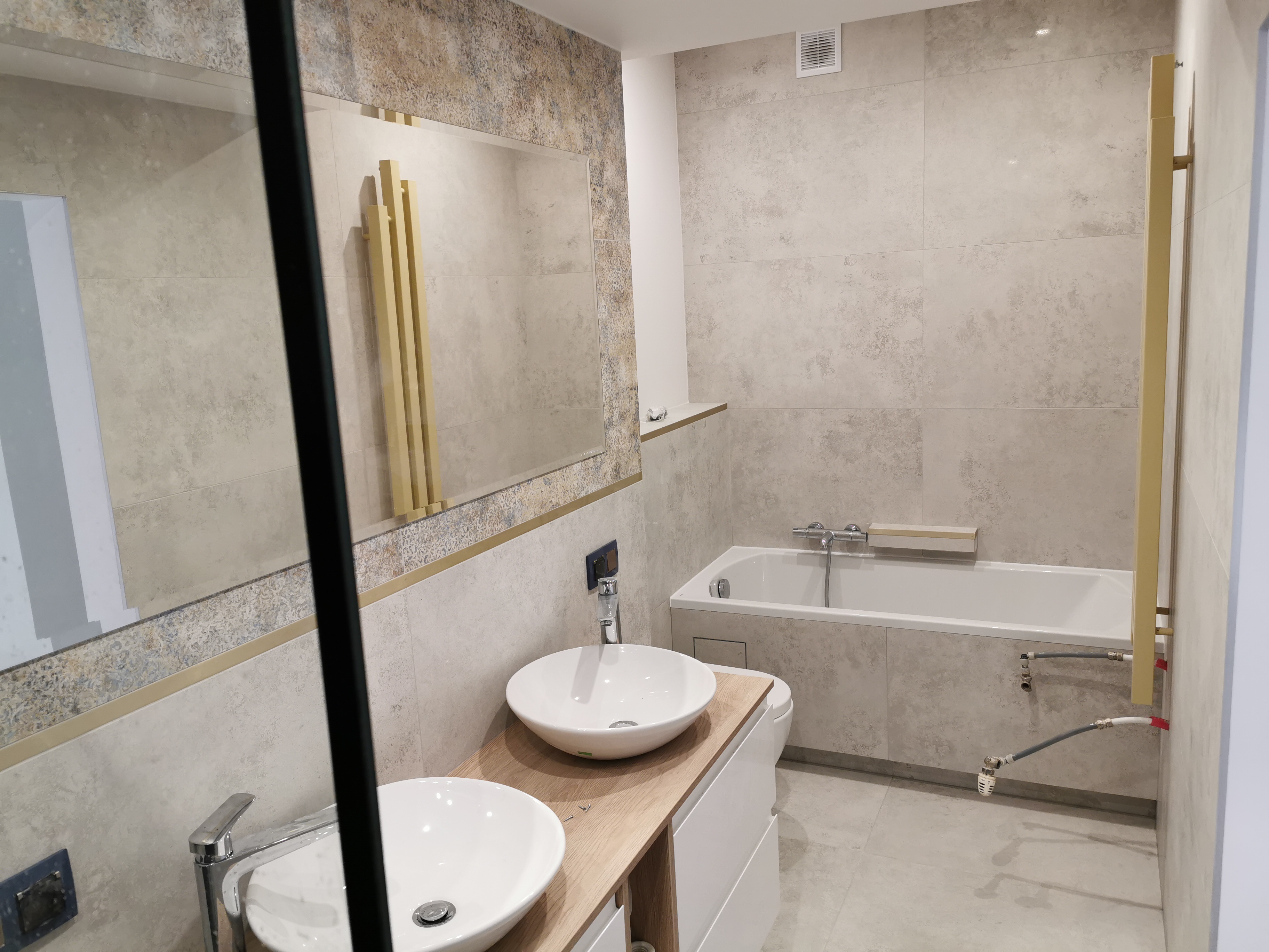 Nowoczesna łazienka z dwoma okrągłymi umywalkami nablatowymi na drewnianym blacie, beżowe płytki na ścianach i podłodze, wanna obudowana płytkami, złote akcenty w postaci grzejnika i ramy lustra.