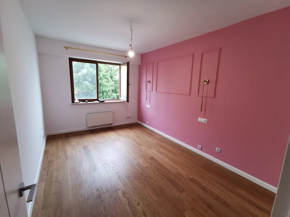 Pusty pokój po odświeżeniu: różowa ściana z dekoracyjnymi ramkami i złotymi lampami ściennymi, drewniana podłoga, biała ściana z oknem i grzejnikiem.