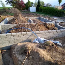 Wznoszenie fundamentów domu jednorodzinnego z bloczków betonowych, widoczne wykopy, hałdy ziemi, niebieski wąż instalacyjny i worki z zaprawą.