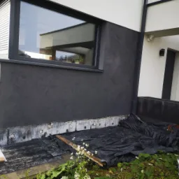 Fragment elewacji budynku z oknem i szarą, strukturalną powłoką, widoczne zabezpieczenia folią i deski na trawie, białe kwiaty na pierwszym planie.